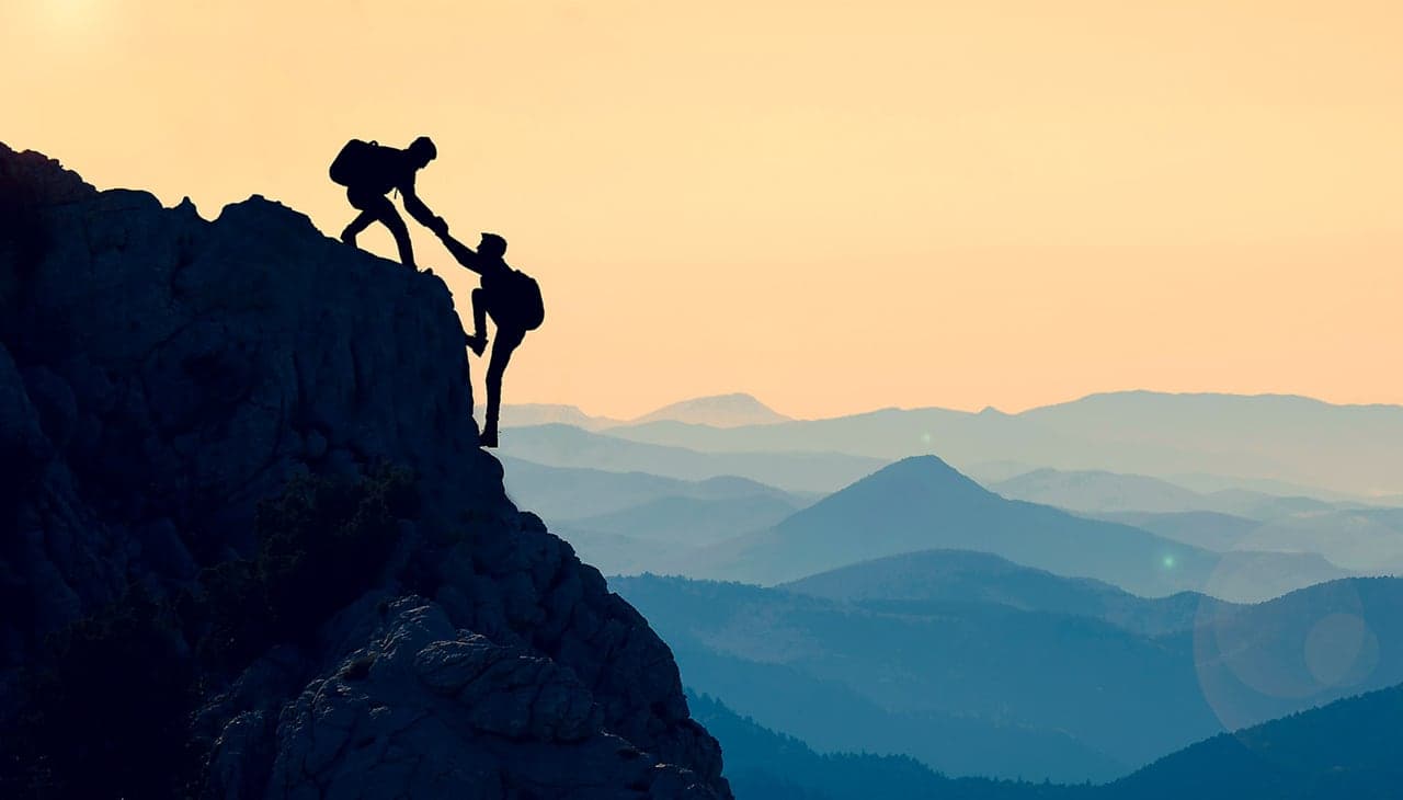 One climber reaching down to help another up the final stretch of a summit